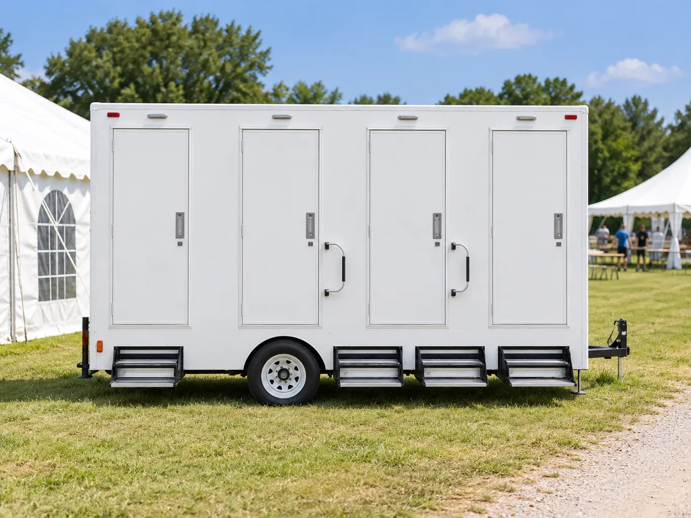 A white four-stall restroom trailer parked on grass between two white event marquees on a clear morning
