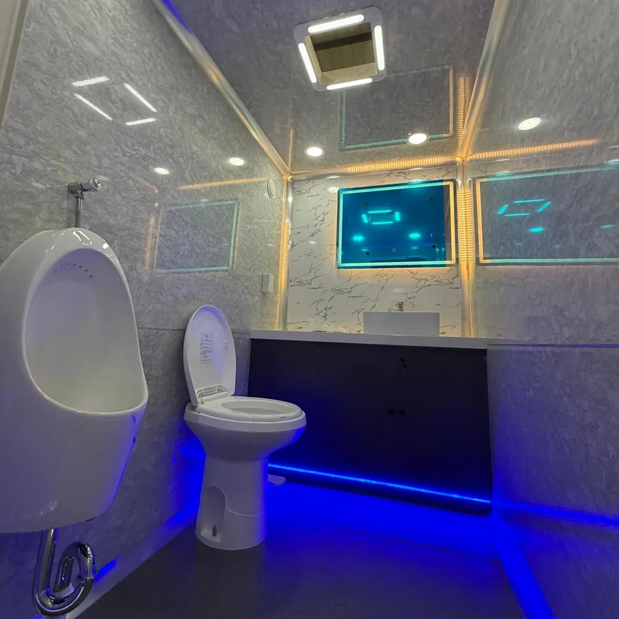Inside a men’s restroom: a urinal, a flush toilet and a vanity with a sink beneath a backlit mirror and a ceiling air-conditioning unit